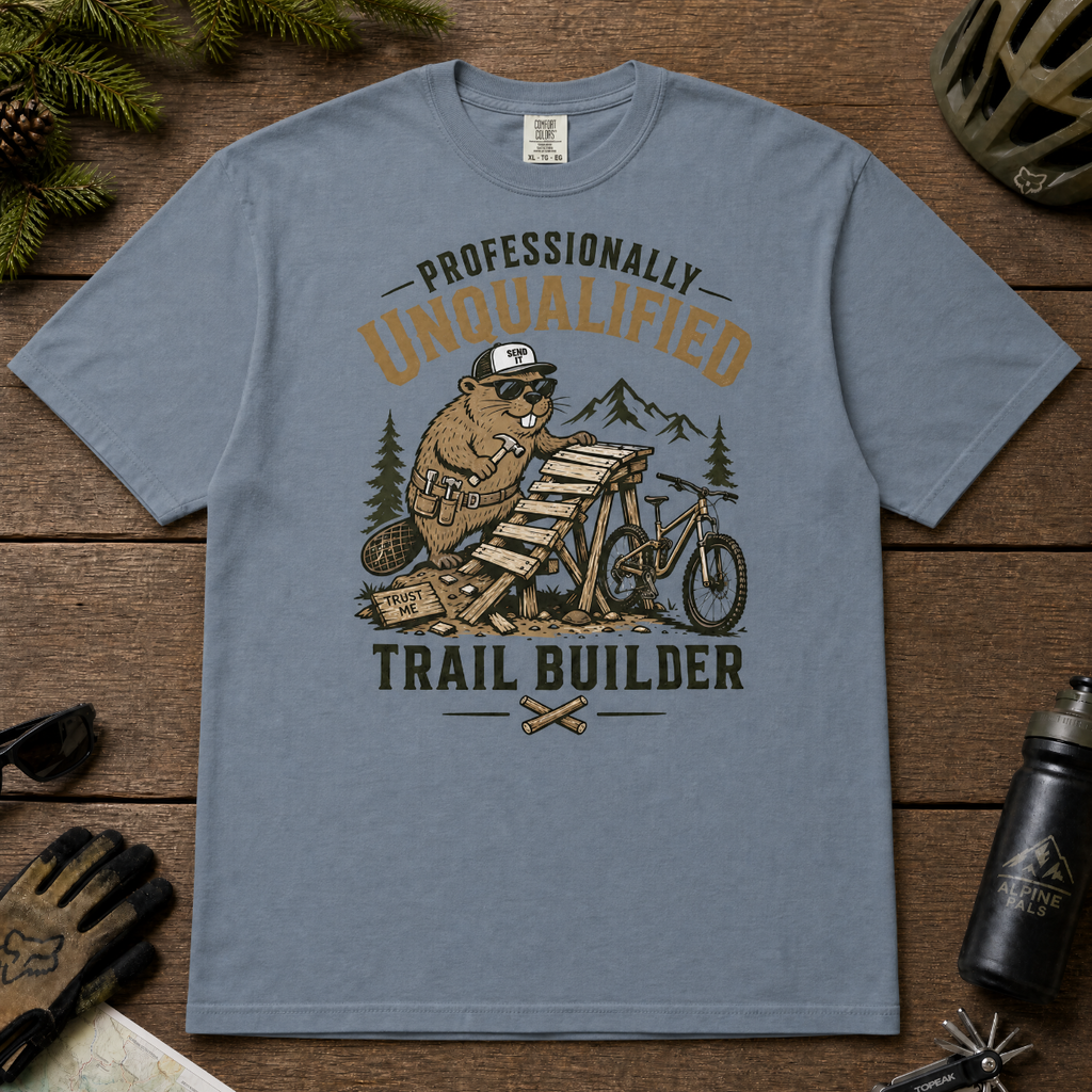 Blue jean colored t-shirt with graphic design of a beaver building a trail, surrounded by outdoor gear on a wooden surface.