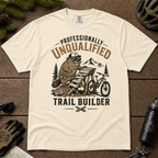 Ivory white t-shirt with a graphic of a beaver building a trail and text 'Professionally Unqualified Trail Builder' on a wooden surface.