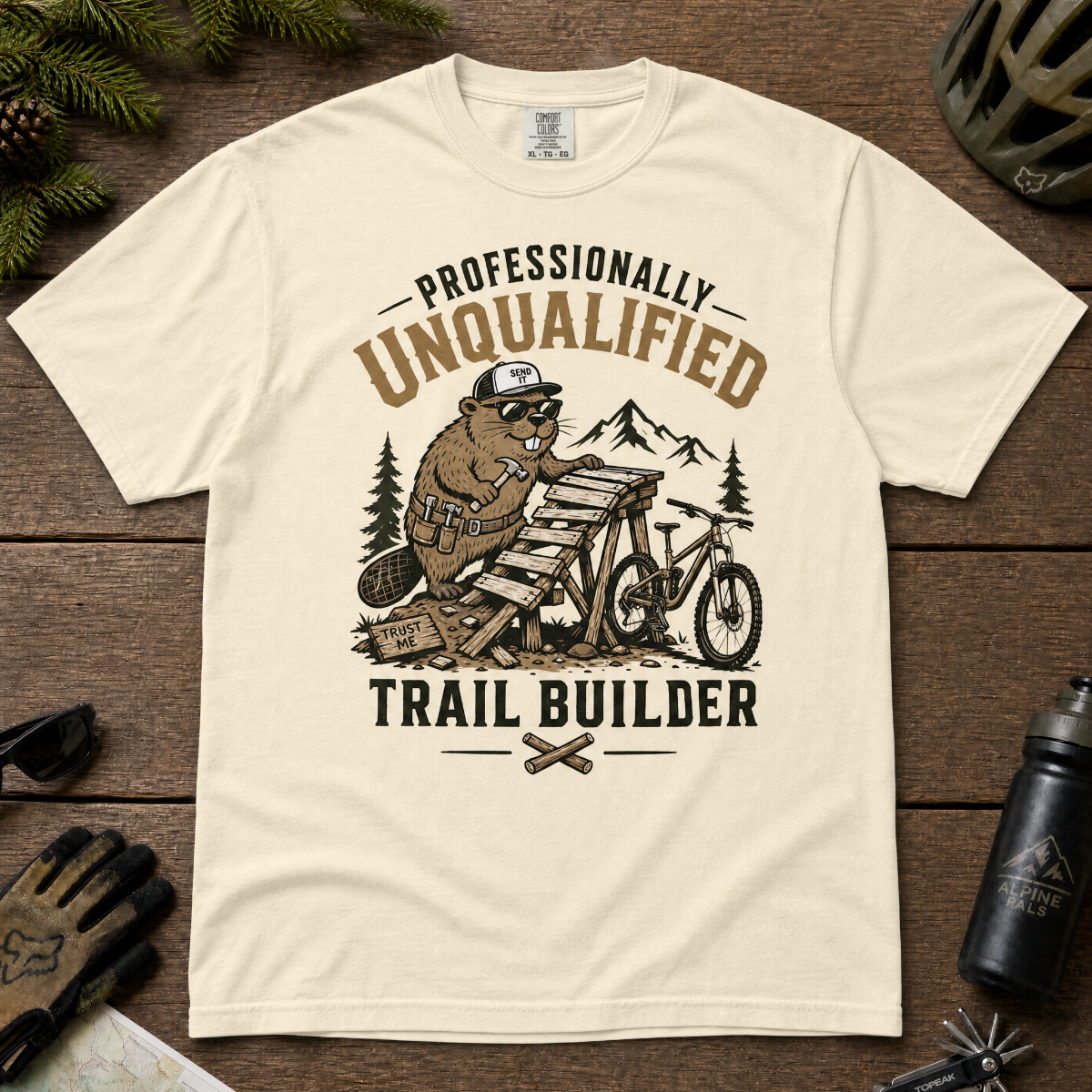 Ivory white t-shirt with a graphic of a beaver building a trail and text 'Professionally Unqualified Trail Builder' on a wooden surface.