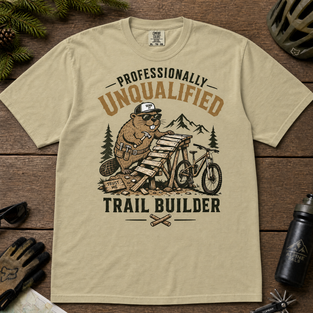 Khaki colored t-shirt with graphic design of a beaver building a trail, surrounded by outdoor gear on a wooden surface.