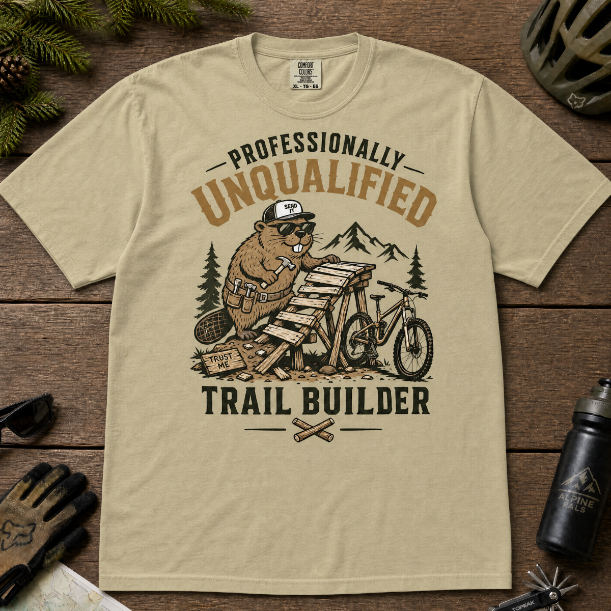 Khaki colored t-shirt with graphic design of a beaver building a trail, surrounded by outdoor gear on a wooden surface.