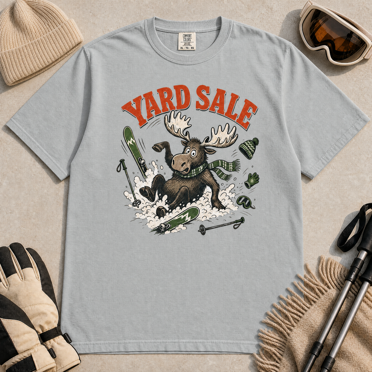 Granite gray t-shirt with moose graphic and 'Yard Sale' text, surrounded by winter accessories on a light background