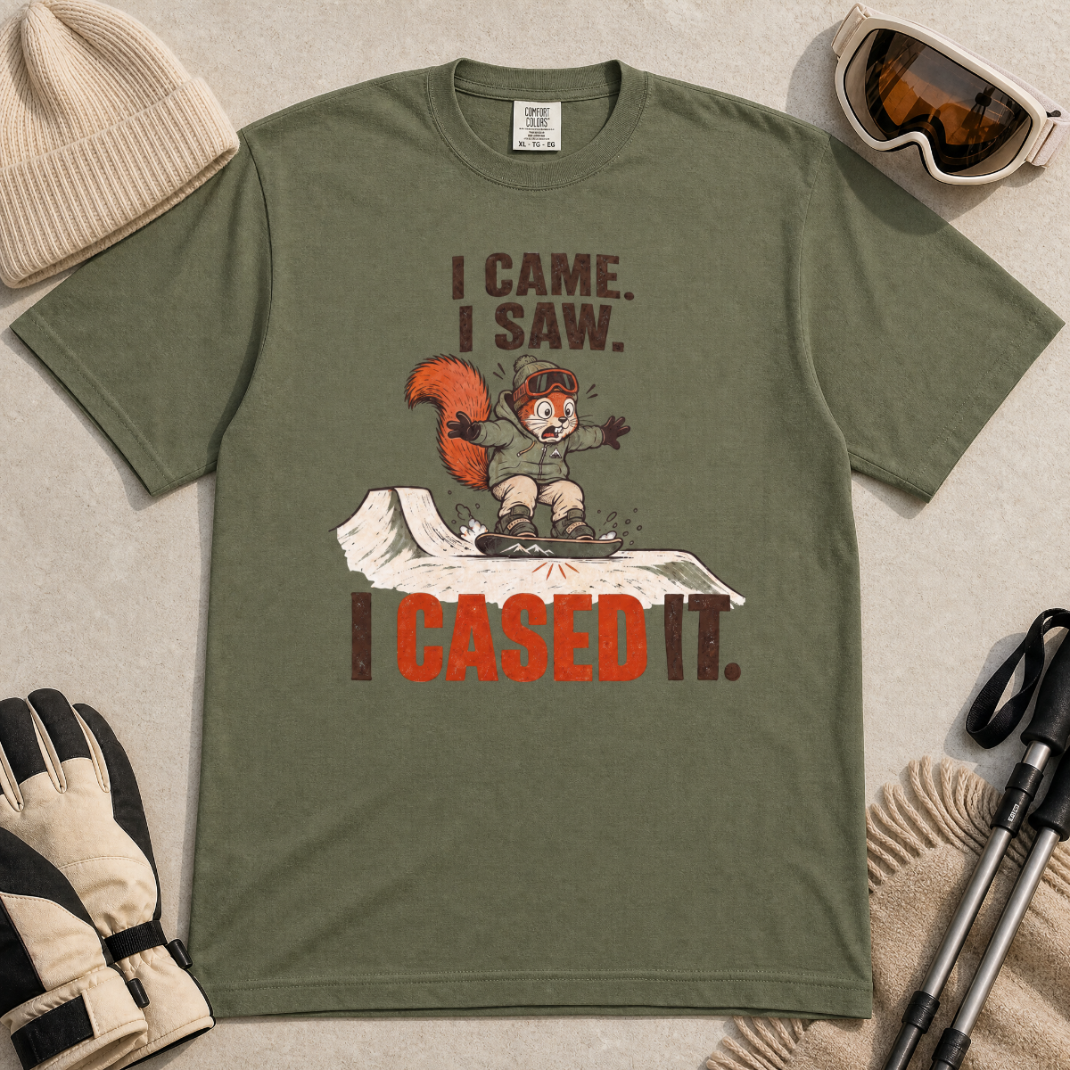 Moss green t-shirt with a graphic design featuring a cartoon character and text, surrounded by winter accessories on a light background.