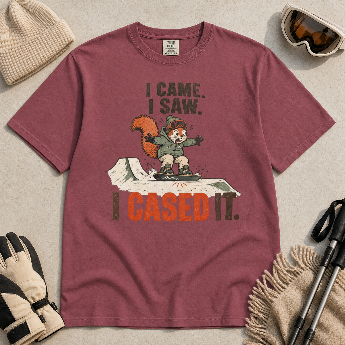 Brick red t-shirt with a graphic design of a squirrel and text on a beige surface with winter accessories.