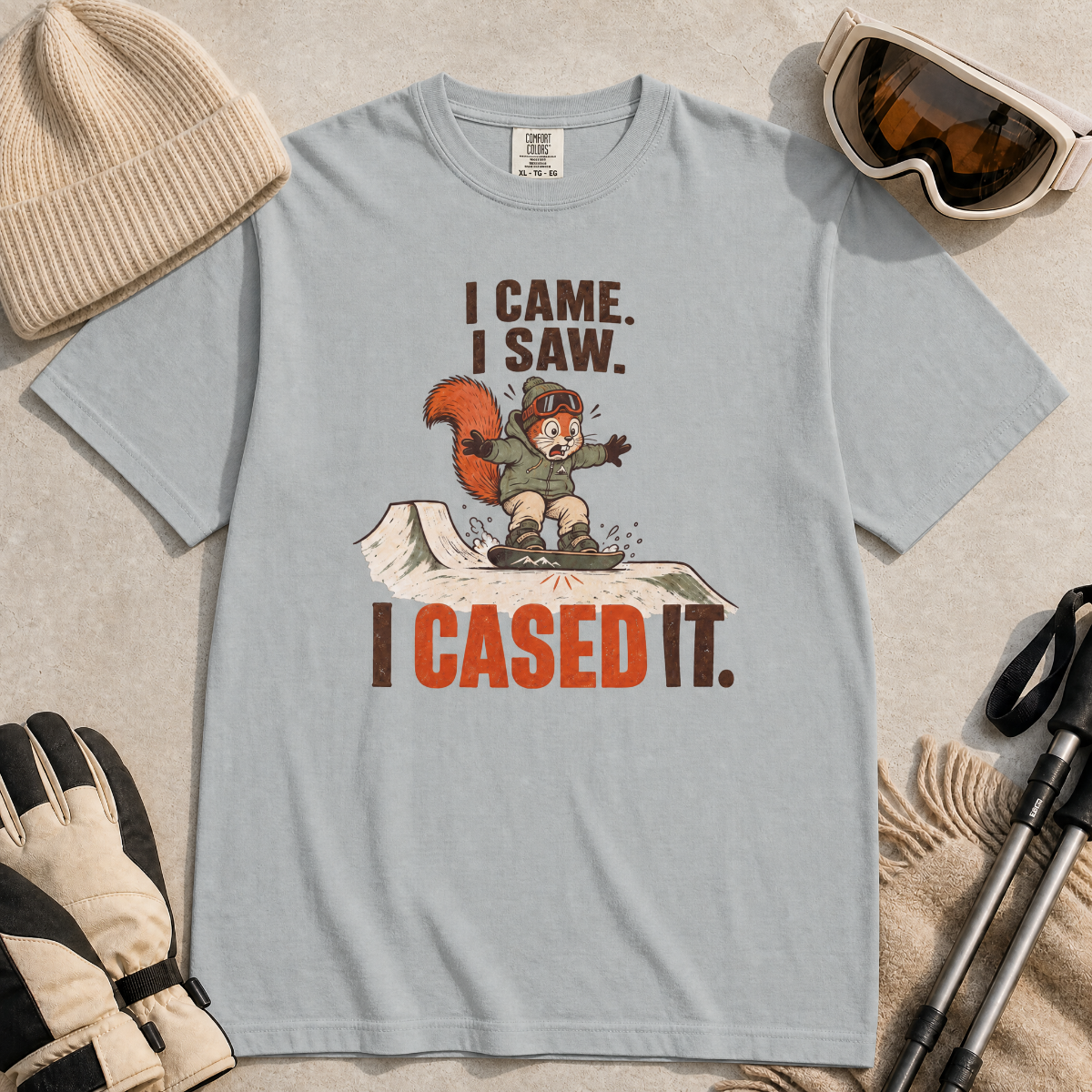 Granite gray t-shirt with a graphic design featuring a squirrel and text, surrounded by winter accessories on a beige background.