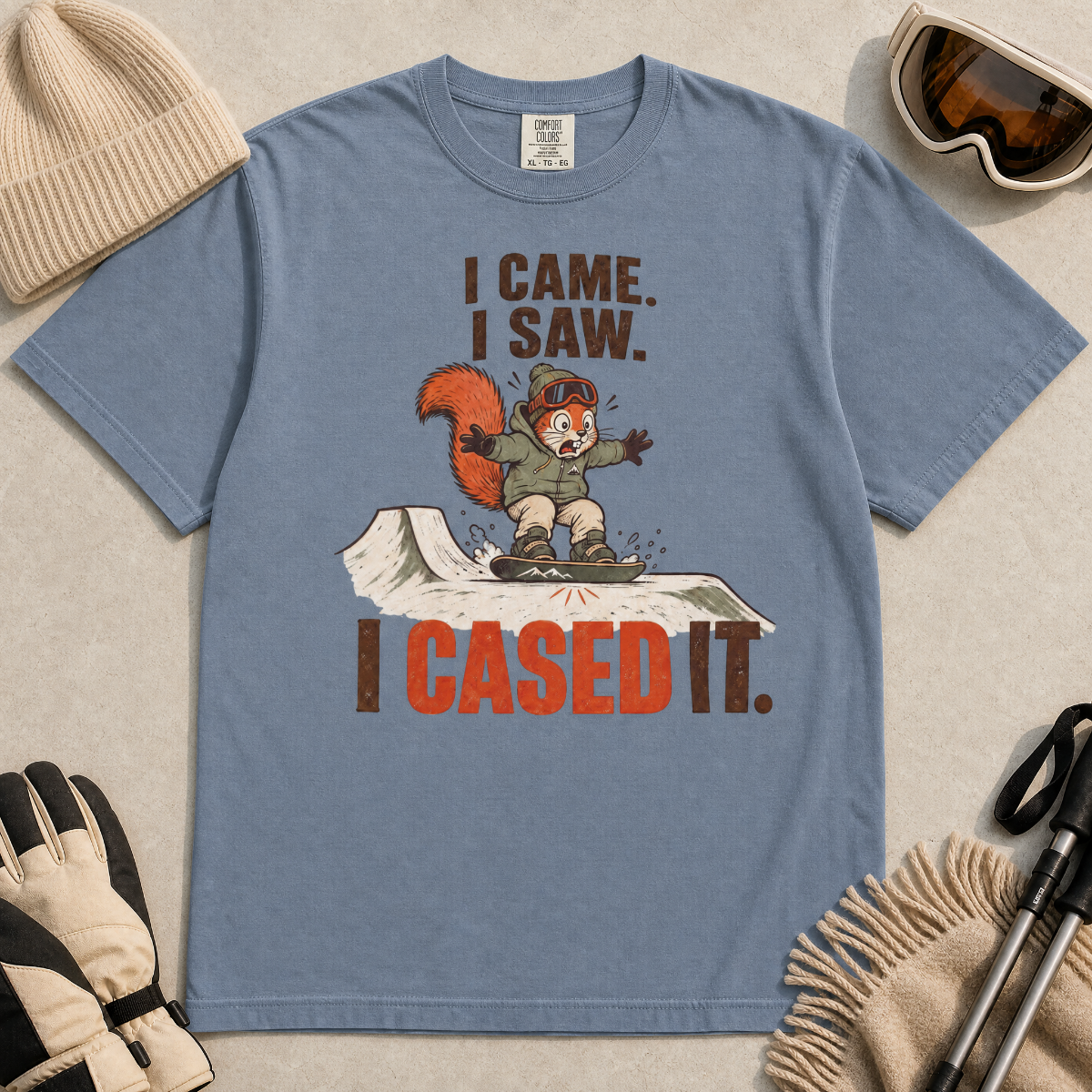 Blue jean colored t-shirt with a graphic of a squirrel on skis and text 'I Came. I Saw. I Cased It.' surrounded by winter accessories.