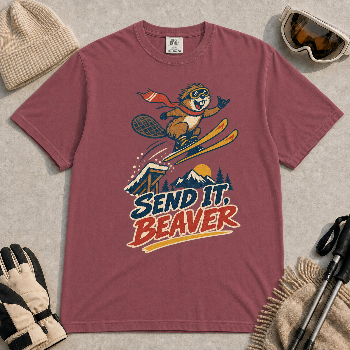 Brick t-shirt featuring a beaver in ski goggles and a flowing scarf launching off a snowy jump above mountain scenery with the text “Send It, Beaver.”