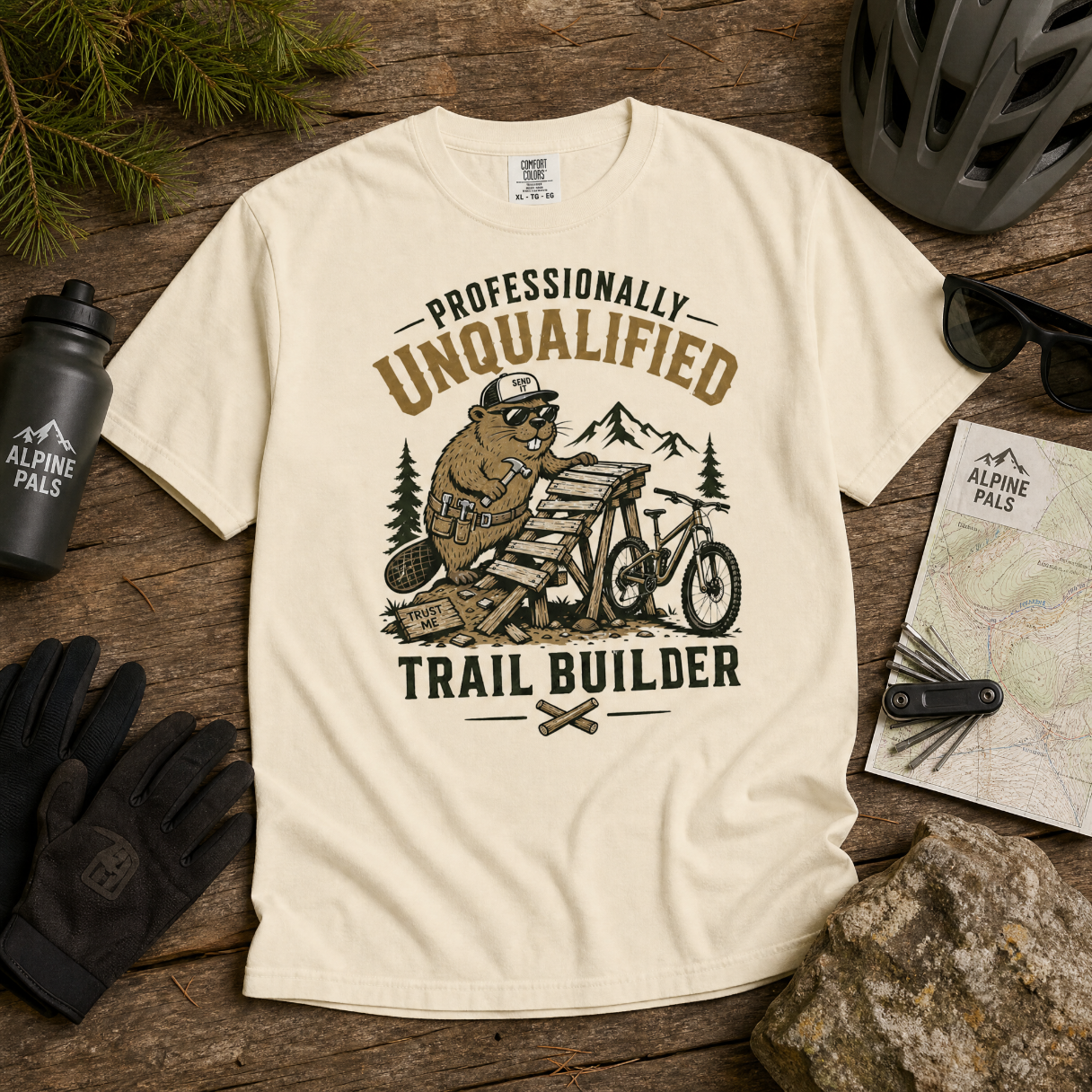 Beige t-shirt with bear graphic and text 'Professionally Unqualified Trail Builder' on a wooden surface with outdoor gear.