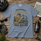 Blue t-shirt with a graphic design of a beaver and text 'Professionally Unqualified Trail Builder' on a wooden surface with outdoor gear.