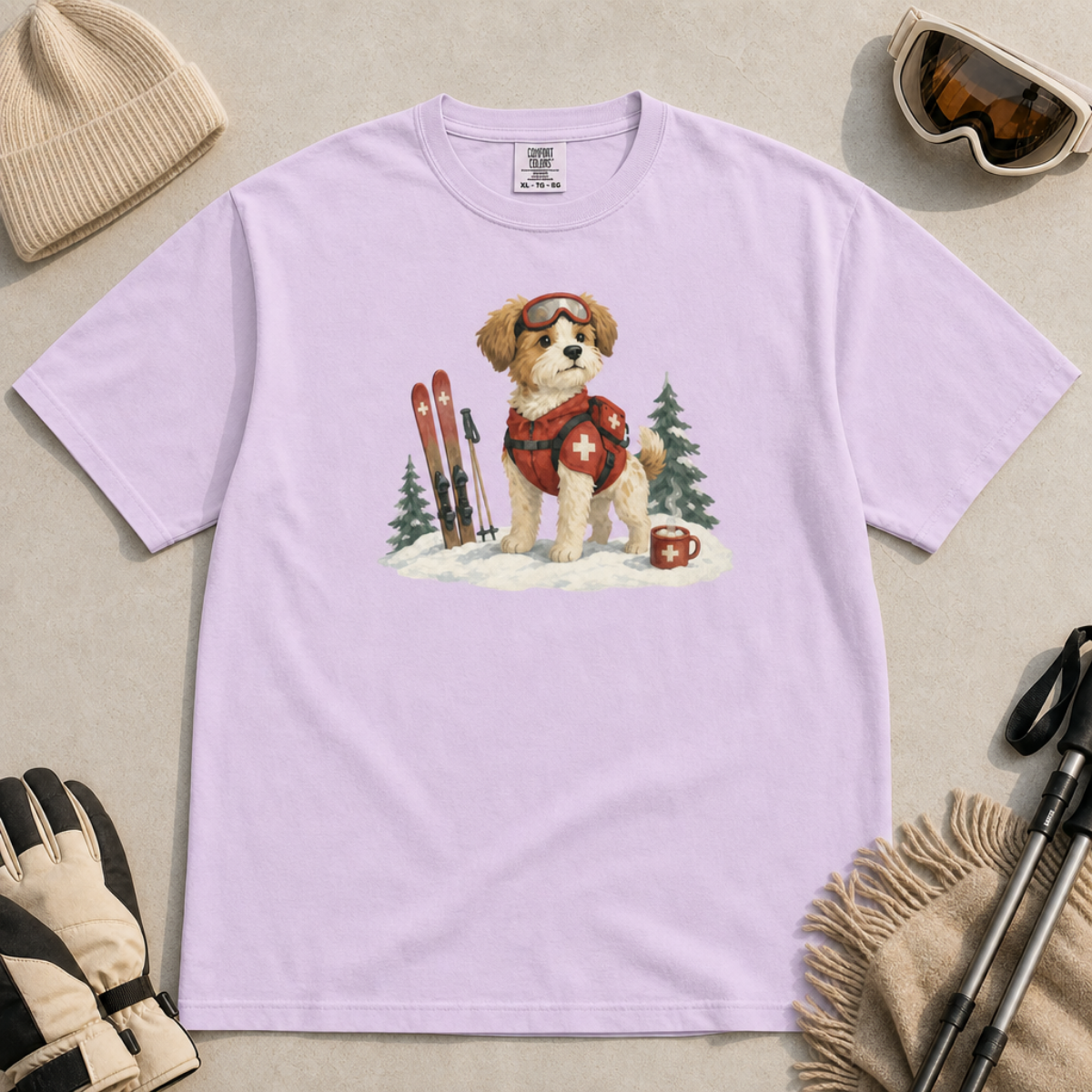 Flat lay of an orchid t-shirt featuring a cute ski patrol dog design with red rescue gear, skis, snowy trees, and a hot cocoa mug.