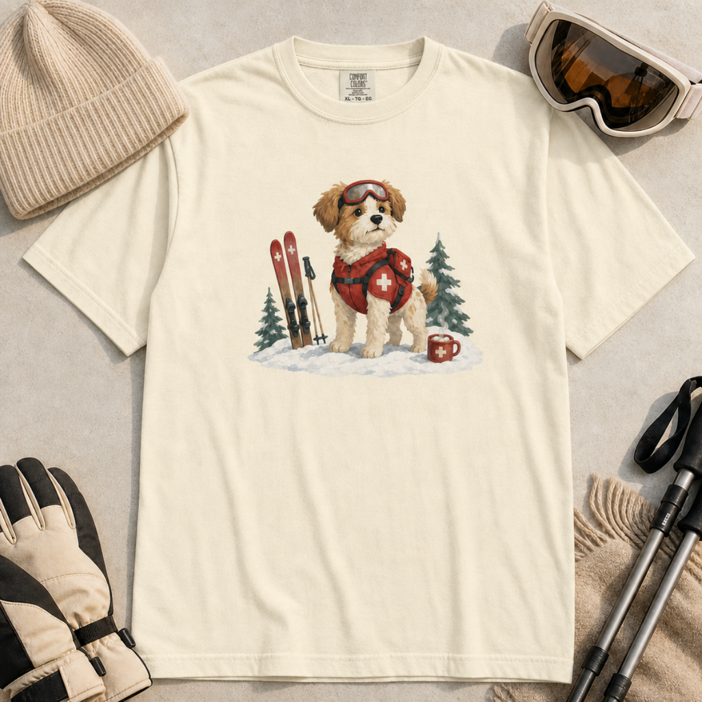 Flat lay of an ivory white t-shirt featuring a cute ski patrol dog design with red rescue gear, skis, snowy trees, and a hot cocoa mug.