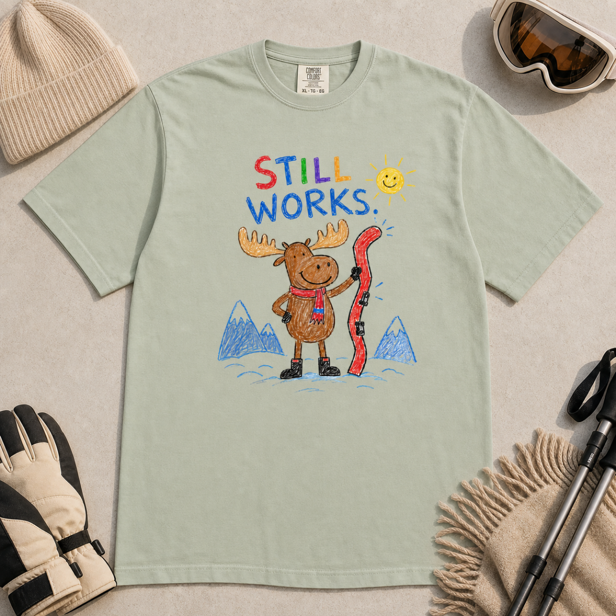 Flat lay of a bay green t-shirt featuring a colourful hand-drawn moose ski graphic with mountains, sun, and “Still Works.” text, styled with winter accessories.