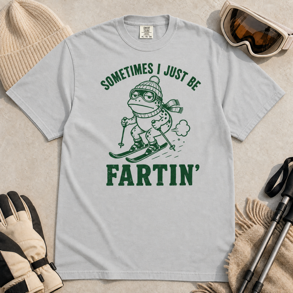 Flat lay of a granite gray t-shirt featuring a funny frog skiing graphic with “Sometimes I Just Be Fartin’” text, styled with winter accessories.