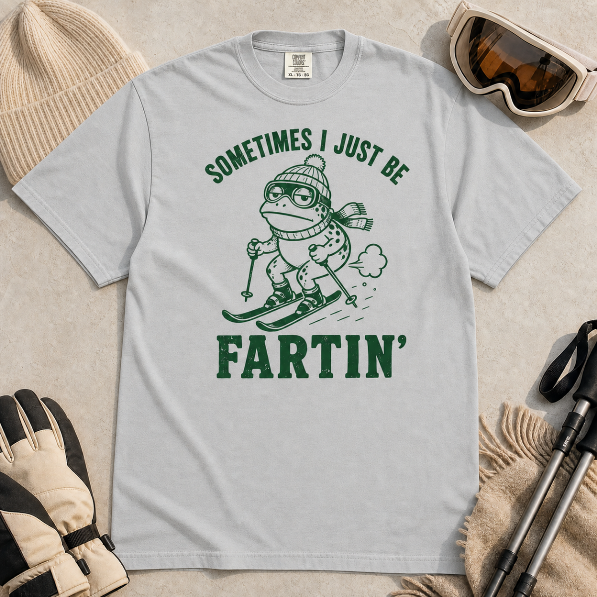 Flat lay of a granite gray t-shirt featuring a funny frog skiing graphic with “Sometimes I Just Be Fartin’” text, styled with winter accessories.