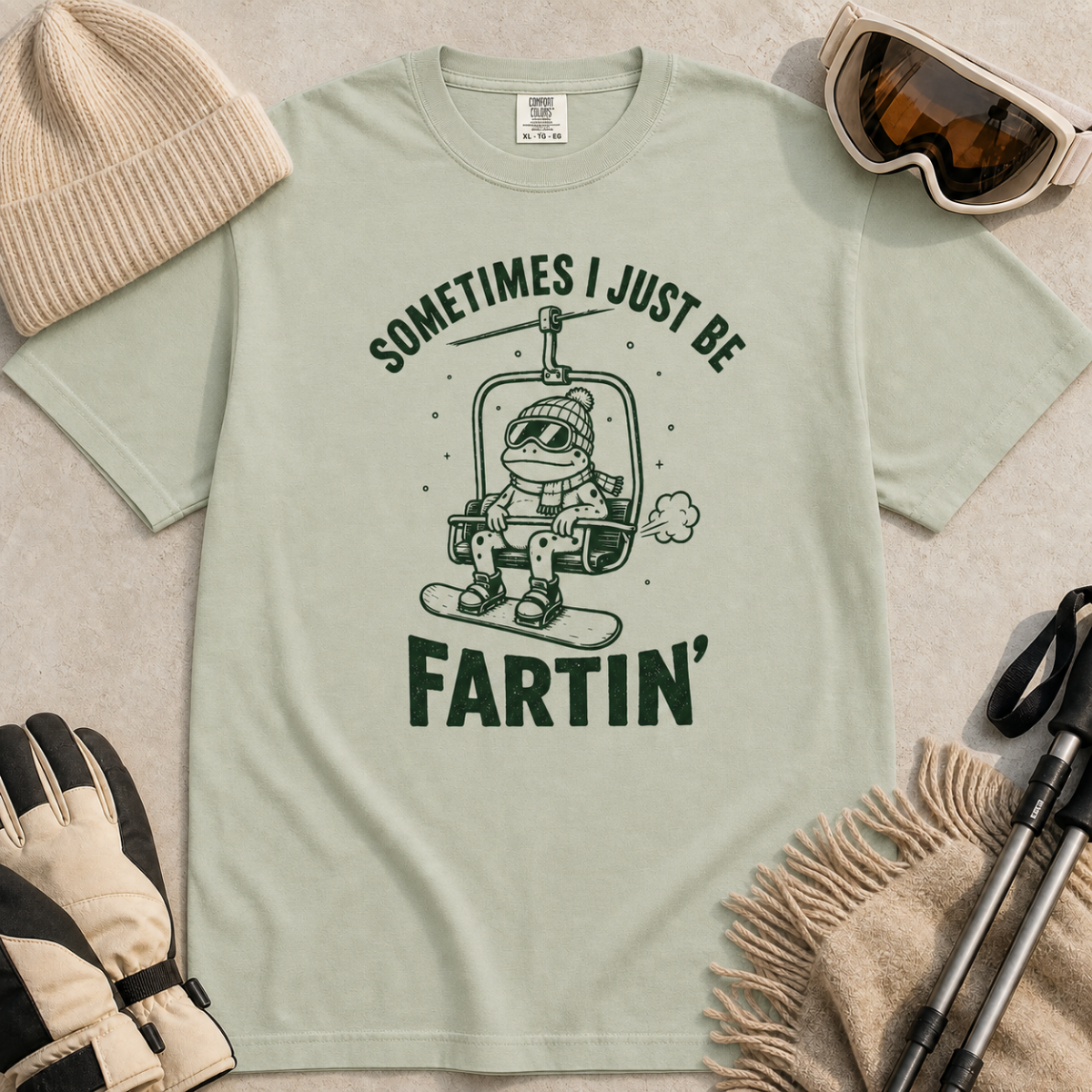 Flat lay of a bay green t-shirt featuring a funny green “Sometimes I Just Be Fartin’” design with a frog riding a chairlift on a snowboard.