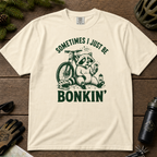 Flat lay of an ivory white t-shirt featuring a “Sometimes I Just Be Bonkin’” raccoon mountain biking graphic, styled with trail accessories on a wooden background.