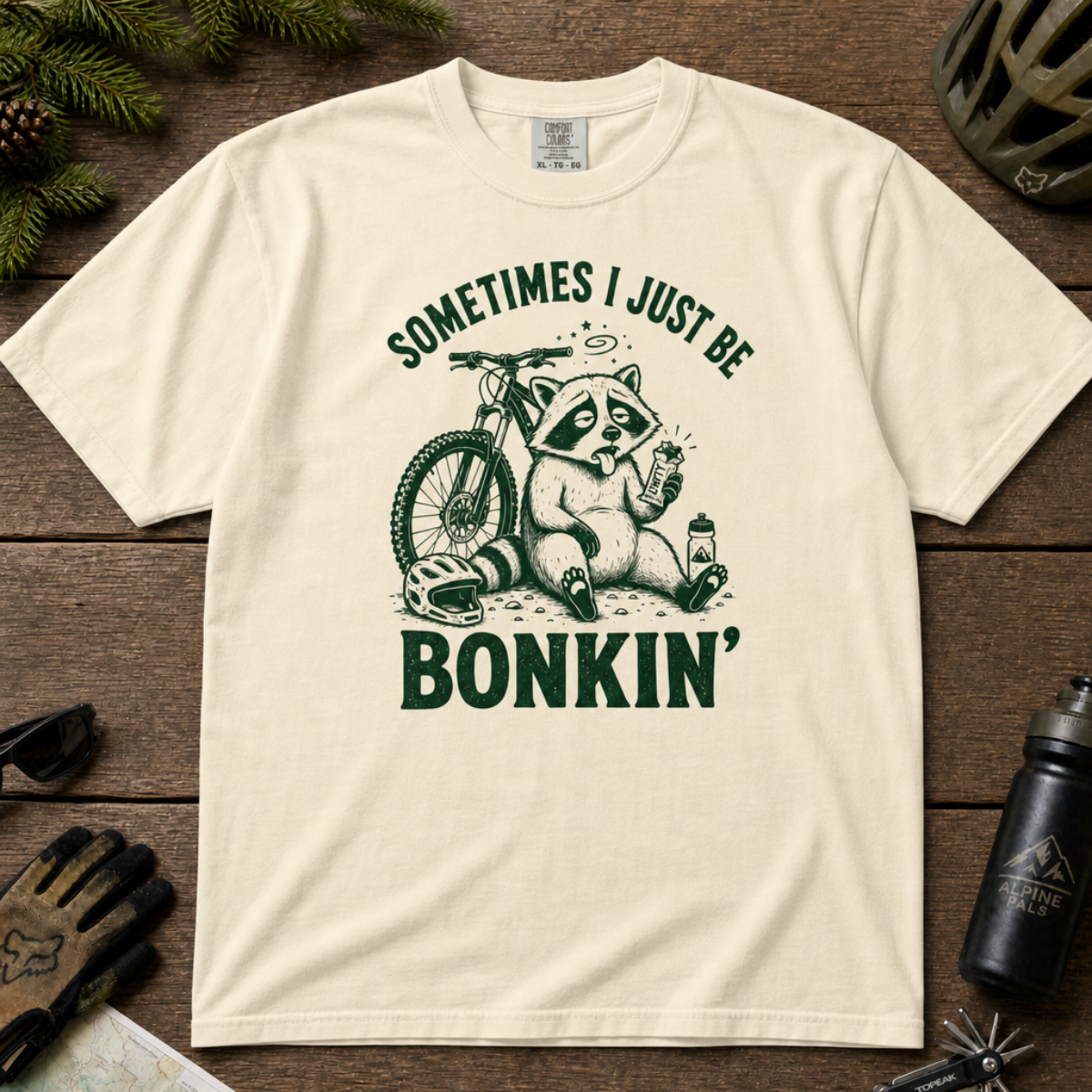 Flat lay of an ivory white t-shirt featuring a “Sometimes I Just Be Bonkin’” raccoon mountain biking graphic, styled with trail accessories on a wooden background.