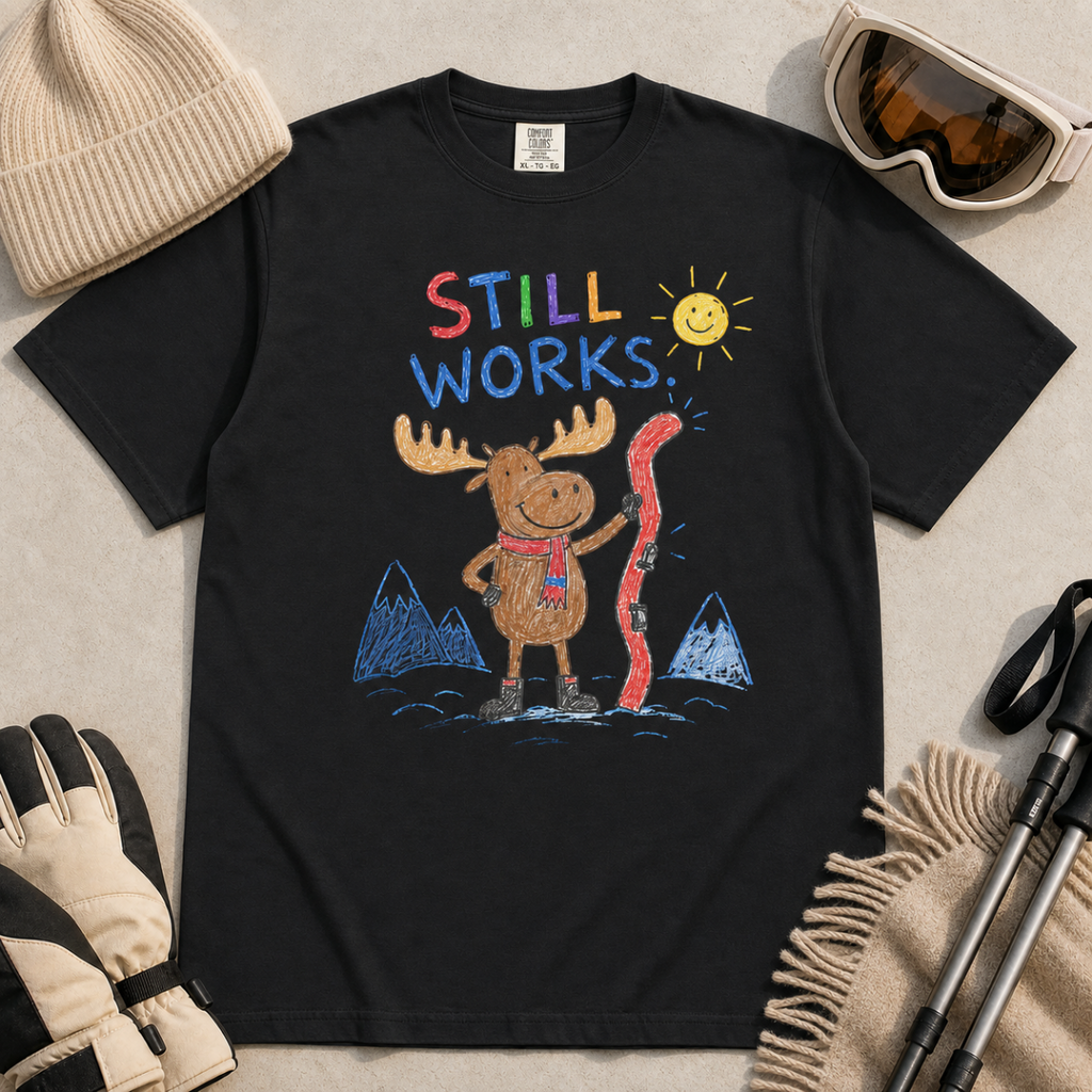 Flat lay of a black t-shirt featuring a colourful hand-drawn moose ski graphic with mountains, sun, and “Still Works.” text, styled with winter accessories.