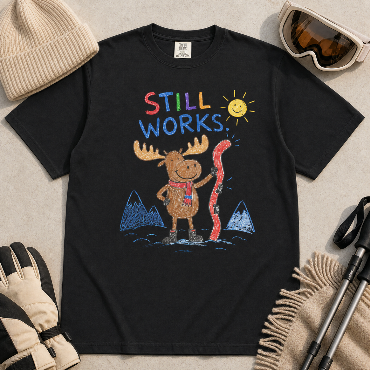 Flat lay of a black t-shirt featuring a colourful hand-drawn moose ski graphic with mountains, sun, and “Still Works.” text, styled with winter accessories.