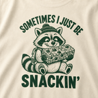 Close-up of an ivory white t-shirt with a playful raccoon snack graphic and bold “Sometimes I Just Be Snackin’” text.