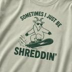 Close-up of a bay green t-shirt with a simple green snowboard goat graphic and bold “Sometimes I Just Be Shreddin’” text.