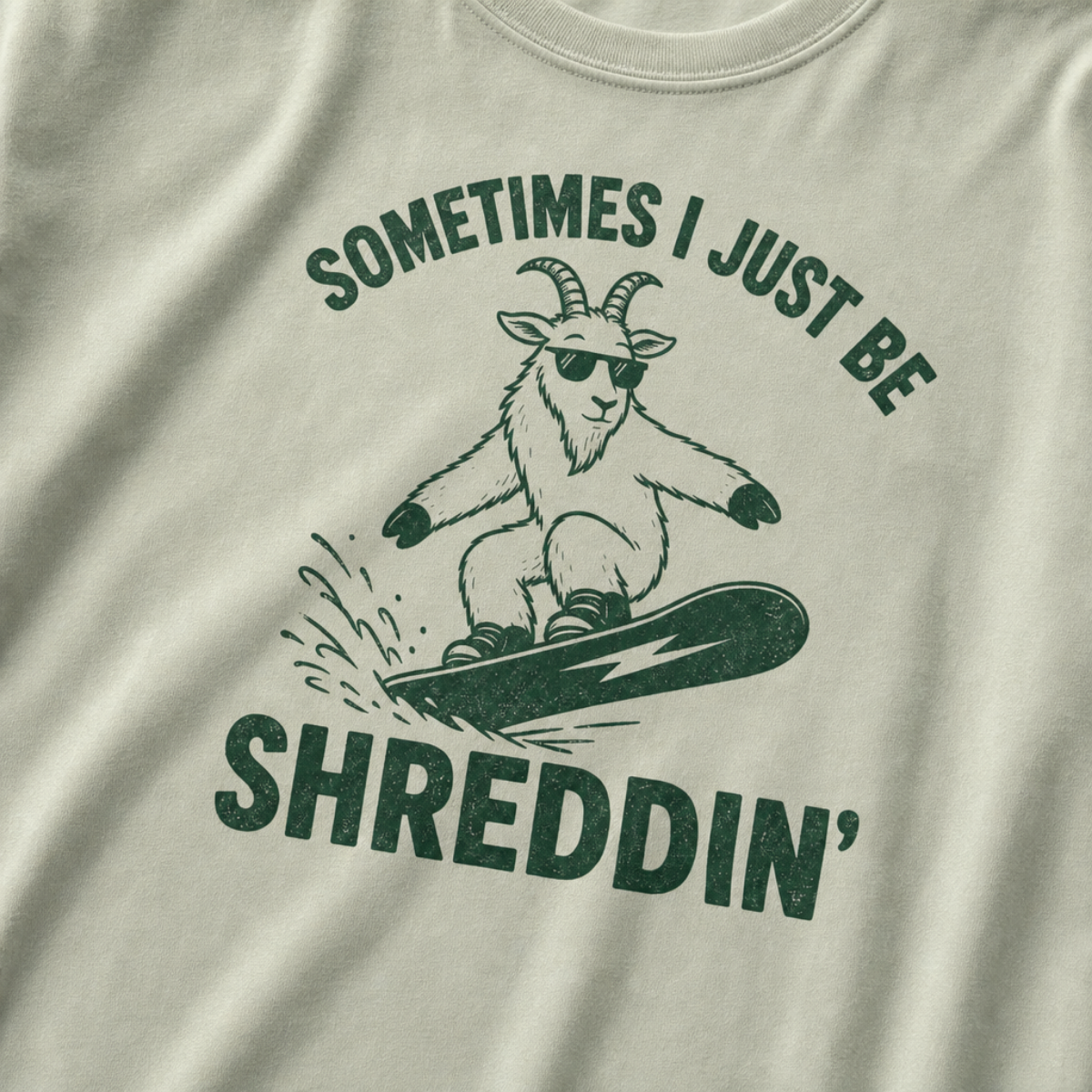 Close-up of a bay green t-shirt with a simple green snowboard goat graphic and bold “Sometimes I Just Be Shreddin’” text.