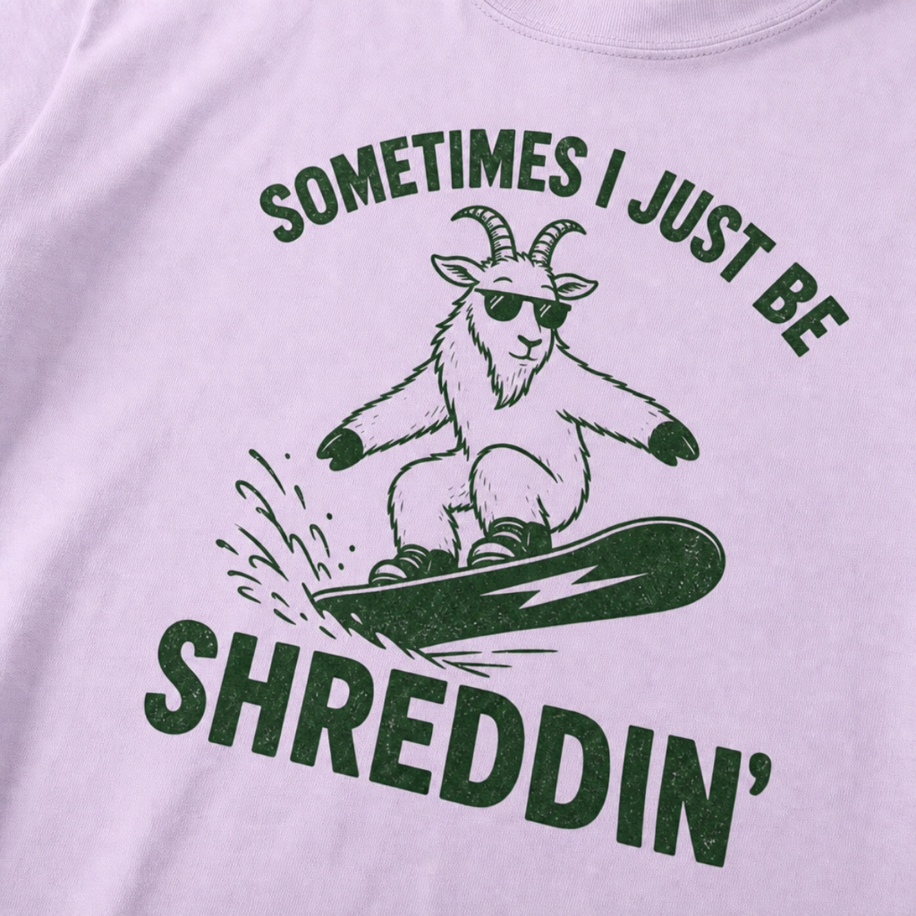 Close-up of an orchid t-shirt with a simple green snowboard goat graphic and bold “Sometimes I Just Be Shreddin’” text.