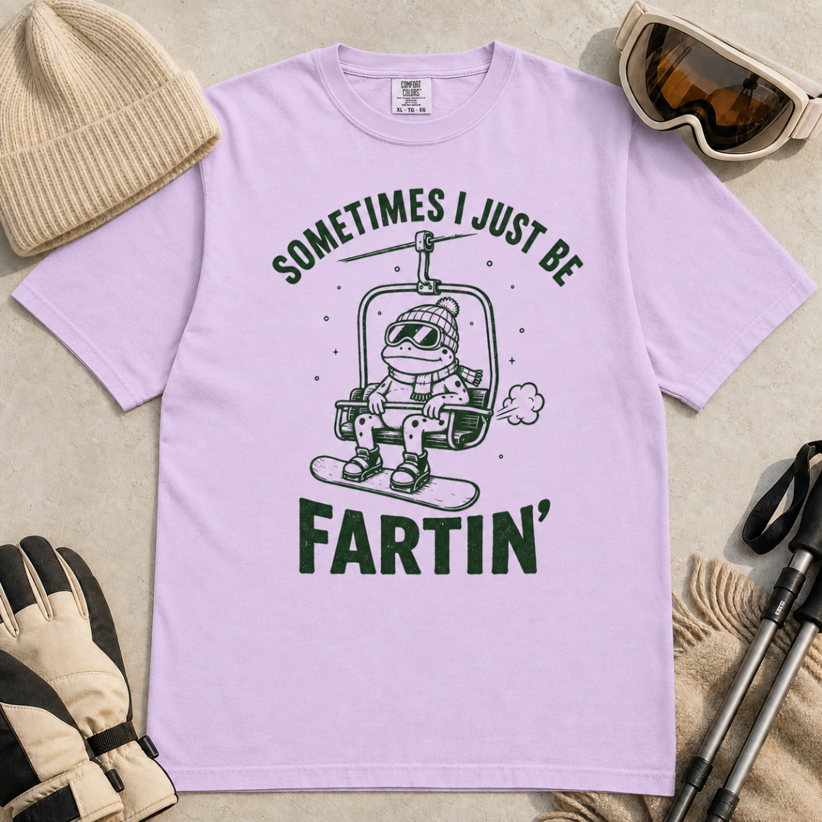 Flat lay of an orchid colored t-shirt featuring a funny green “Sometimes I Just Be Fartin’” design with a frog riding a chairlift on a snowboard.