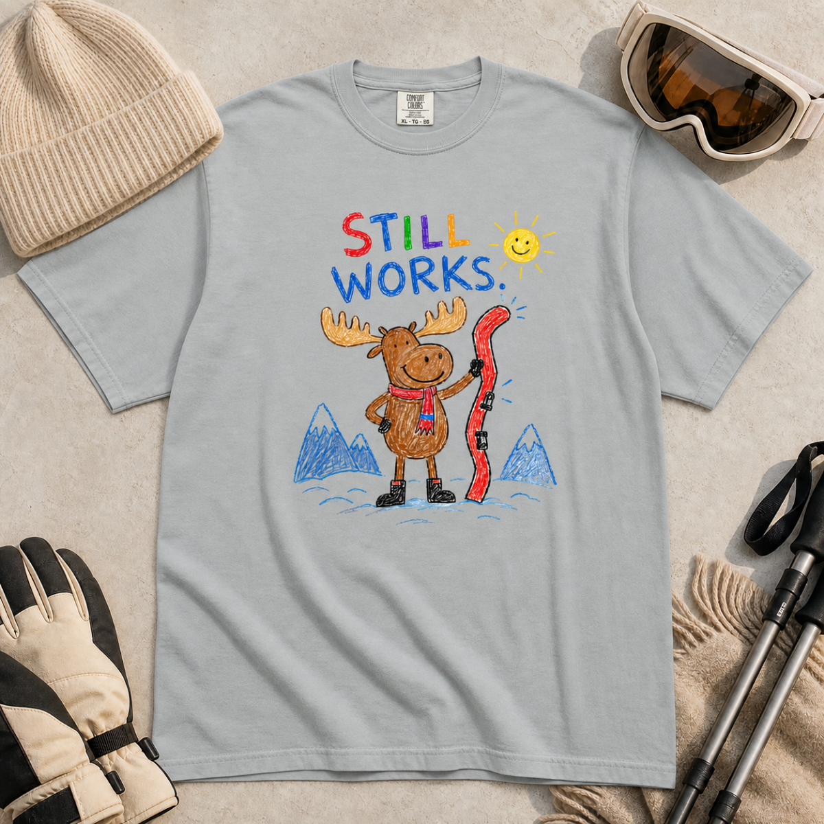 Flat lay of a granite gray t-shirt featuring a colourful hand-drawn moose ski graphic with mountains, sun, and “Still Works.” text, styled with winter accessories.