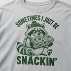 Close-up of a granite gray t-shirt with a playful raccoon snack graphic and bold “Sometimes I Just Be Snackin’” text.