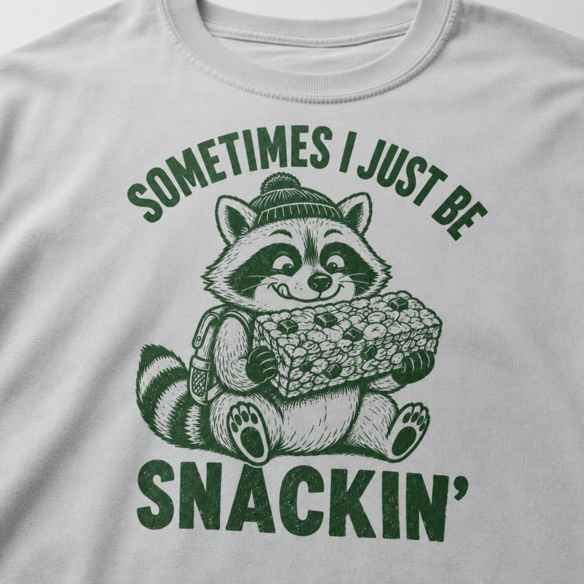 Close-up of a granite gray t-shirt with a playful raccoon snack graphic and bold “Sometimes I Just Be Snackin’” text.