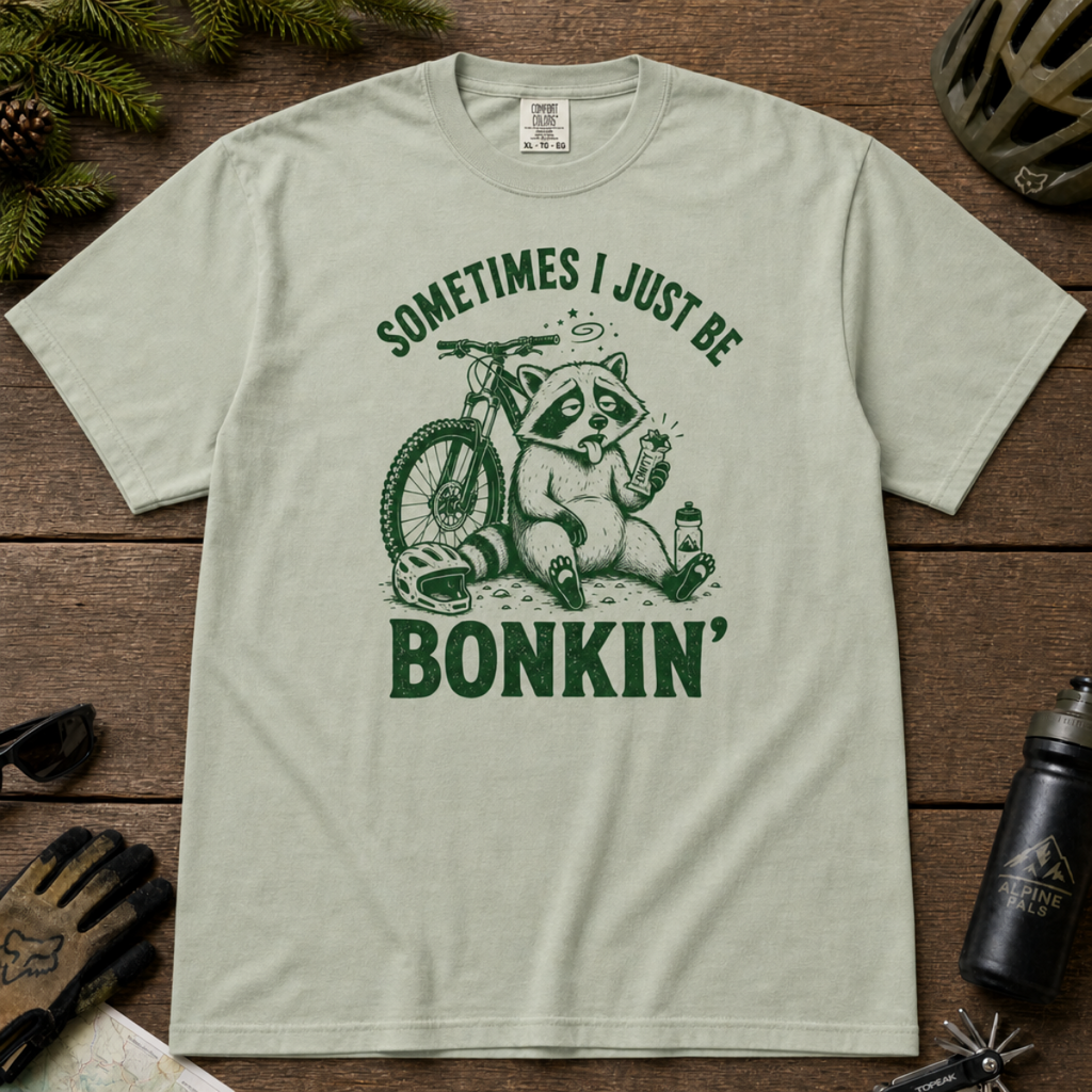 Flat lay of a bay green t-shirt featuring a “Sometimes I Just Be Bonkin’” raccoon mountain biking graphic, styled with trail accessories on a wooden background.