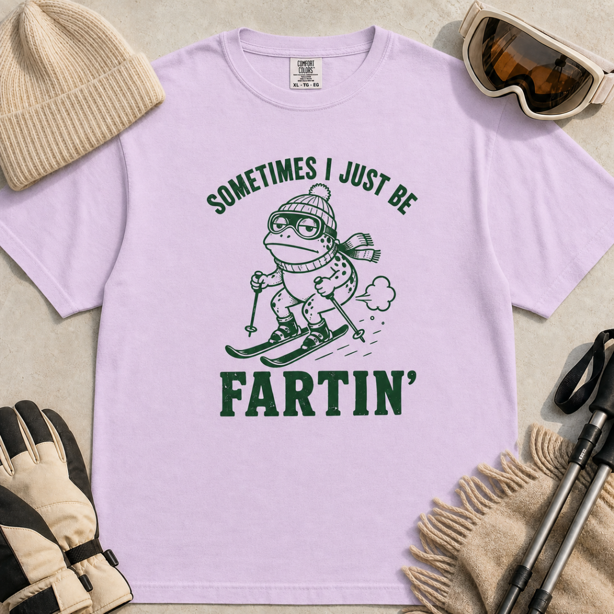 Flat lay of an orchid colored  t-shirt featuring a funny frog skiing graphic with “Sometimes I Just Be Fartin’” text, styled with winter accessories.