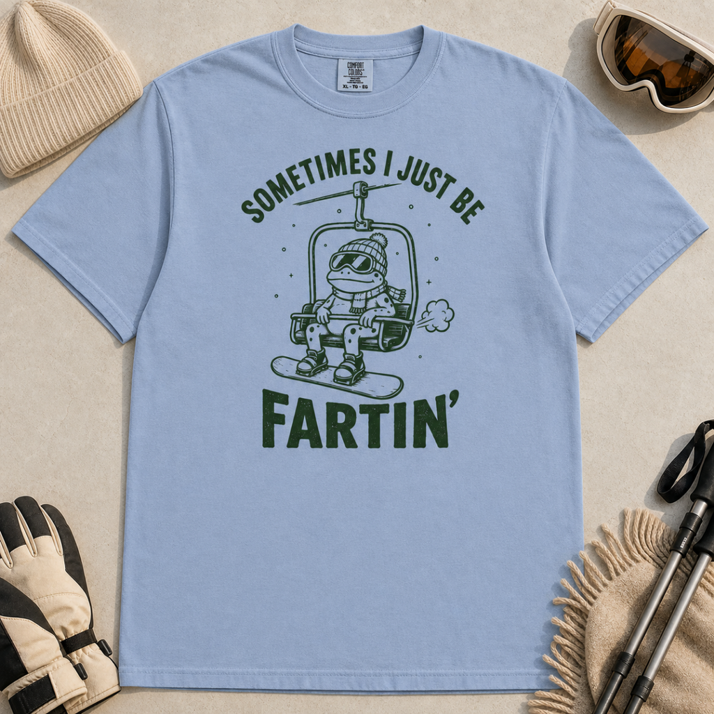 Flat lay of a washed denim blue t-shirt featuring a funny green “Sometimes I Just Be Fartin’” design with a frog riding a chairlift on a snowboard.