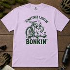 Flat lay of an orchid t-shirt featuring a “Sometimes I Just Be Bonkin’” raccoon mountain biking graphic, styled with trail accessories on a wooden background.