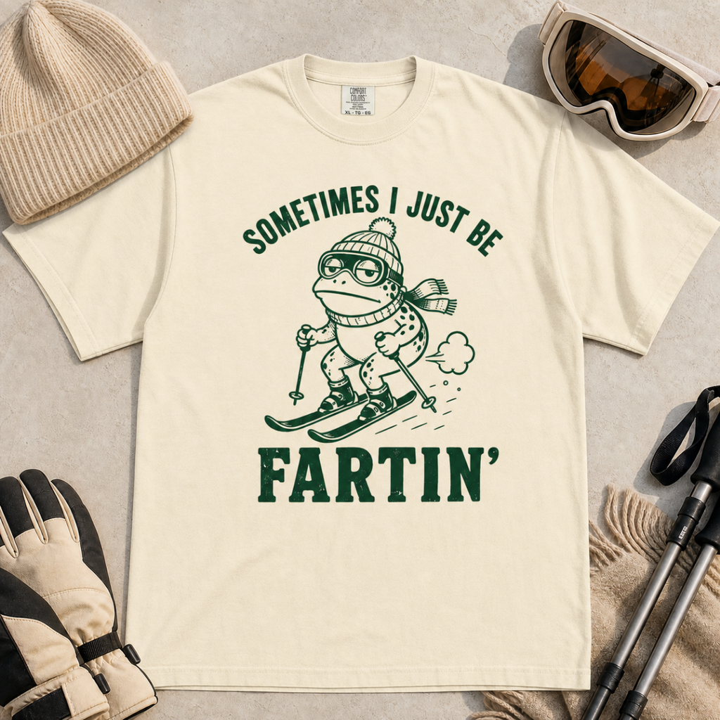 Flat lay of an ivory white t-shirt featuring a funny frog skiing graphic with “Sometimes I Just Be Fartin’” text, styled with winter accessories.