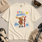 Flat lay of an ivory white t-shirt featuring a colourful hand-drawn moose ski graphic with mountains, sun, and “Still Works.” text, styled with winter accessories.
