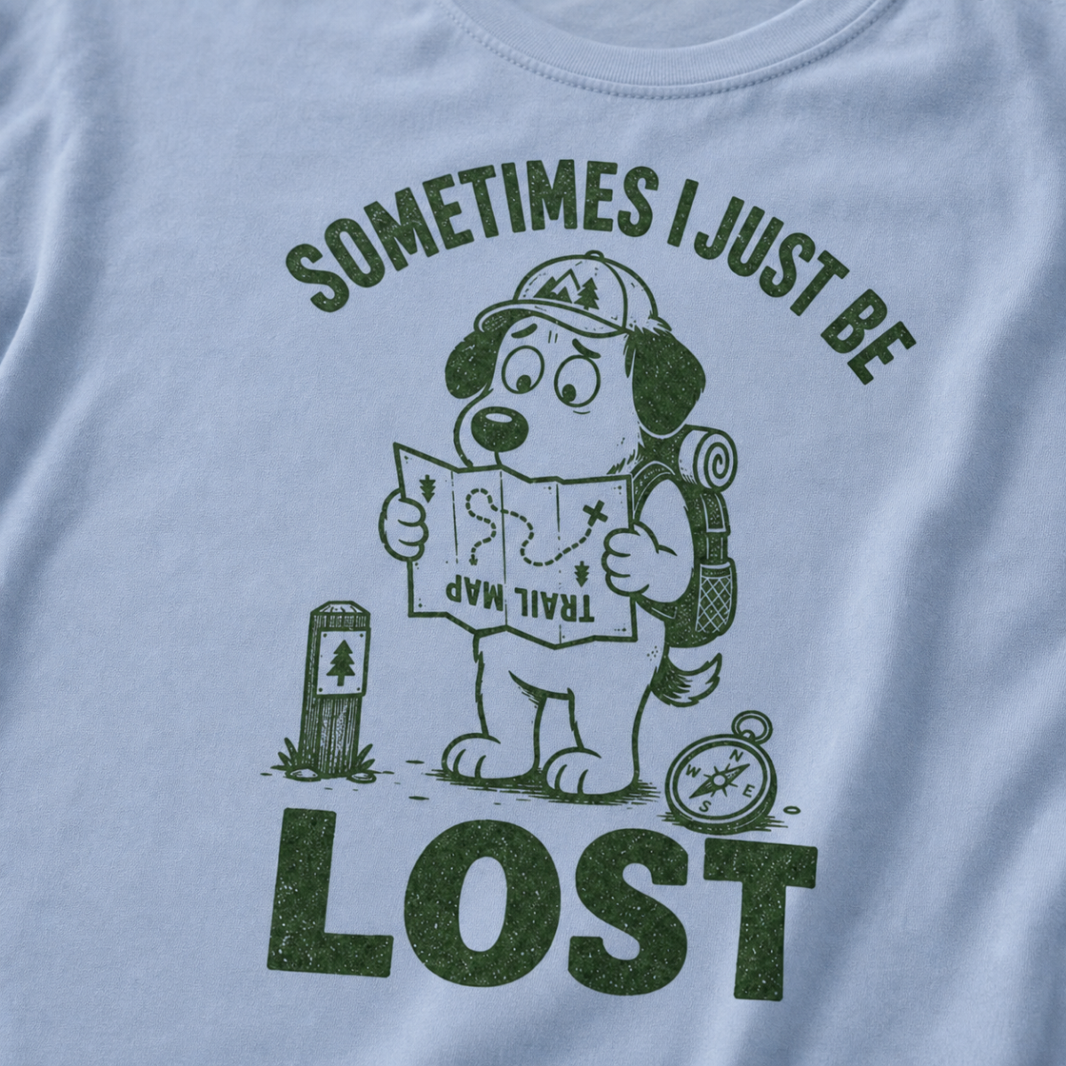Close-up of a WASHED DENIM BLUE t-shirt with a green “Sometimes I Just Be Lost” trail design showing a confused dog reading an upside-down map.
