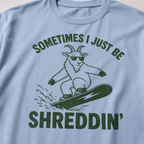 Close-up of a washed denim blue t-shirt with a simple green snowboard goat graphic and bold “Sometimes I Just Be Shreddin’” text.