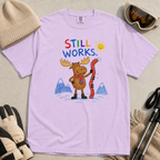 Flat lay of an orchid t-shirt featuring a colourful hand-drawn moose ski graphic with mountains, sun, and “Still Works.” text, styled with winter accessories.