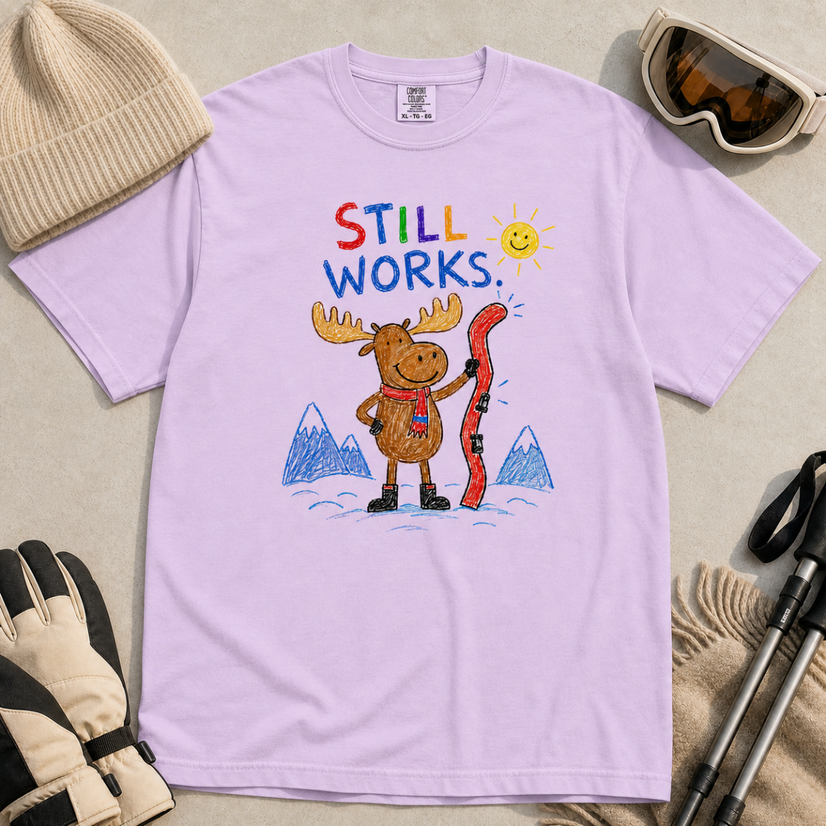 Flat lay of an orchid t-shirt featuring a colourful hand-drawn moose ski graphic with mountains, sun, and “Still Works.” text, styled with winter accessories.