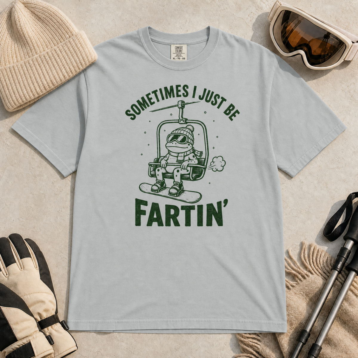 Flat lay of a granite gray t-shirt featuring a funny green “Sometimes I Just Be Fartin’” design with a frog riding a chairlift on a snowboard.