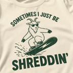 Close-up of an ivory white t-shirt with a simple green snowboard goat graphic and bold “Sometimes I Just Be Shreddin’” text.
