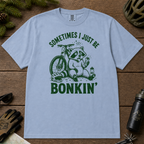 Flat lay of a washed denim blue t-shirt featuring a “Sometimes I Just Be Bonkin’” raccoon mountain biking graphic, styled with trail accessories on a wooden background.