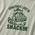 Close-up of a bay green t-shirt with a playful raccoon snack graphic and bold “Sometimes I Just Be Snackin’” text.