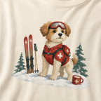 Close-up of an ivory white t-shirt with a sweet ski patrol dog graphic showing a small dog in red rescue gear beside skis and a hot cocoa mug.
