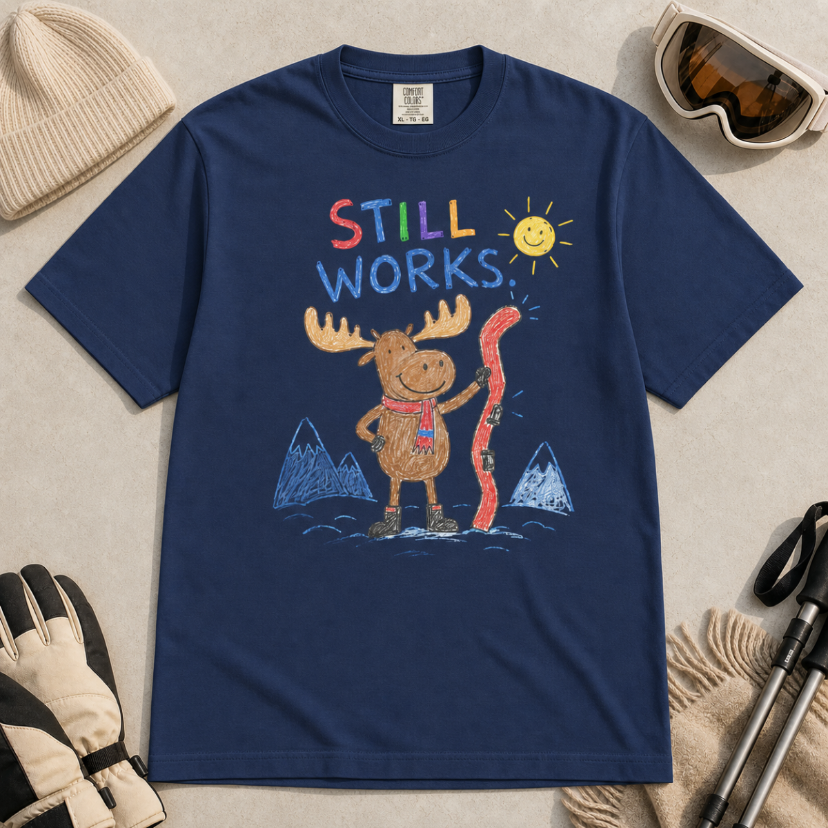 Flat lay of a navy t-shirt featuring a colourful hand-drawn moose ski graphic with mountains, sun, and “Still Works.” text, styled with winter accessories.