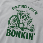 Close-up of a granite gray t-shirt with a funny green mountain biking design showing a tired raccoon bonking beside a bike, helmet, water bottle, and snack bar.