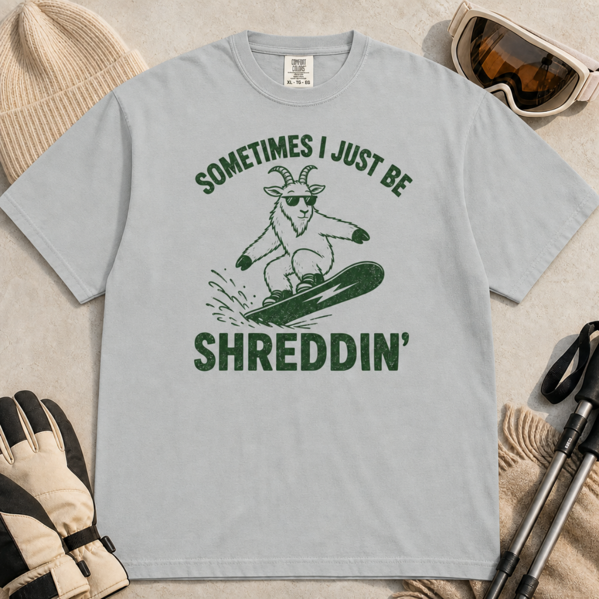Flat lay of a granite gray t-shirt featuring a green “Sometimes I Just Be Shreddin’” design with a goat snowboarding in sunglasses.