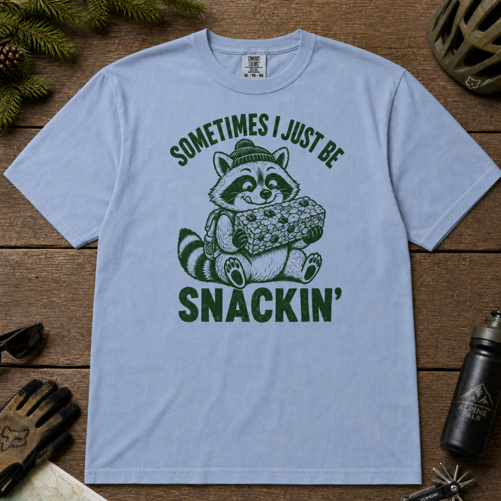 Flat lay of a washed denim blue t-shirt featuring a funny green “Sometimes I Just Be Snackin’” design with a happy raccoon holding a large snack bar.